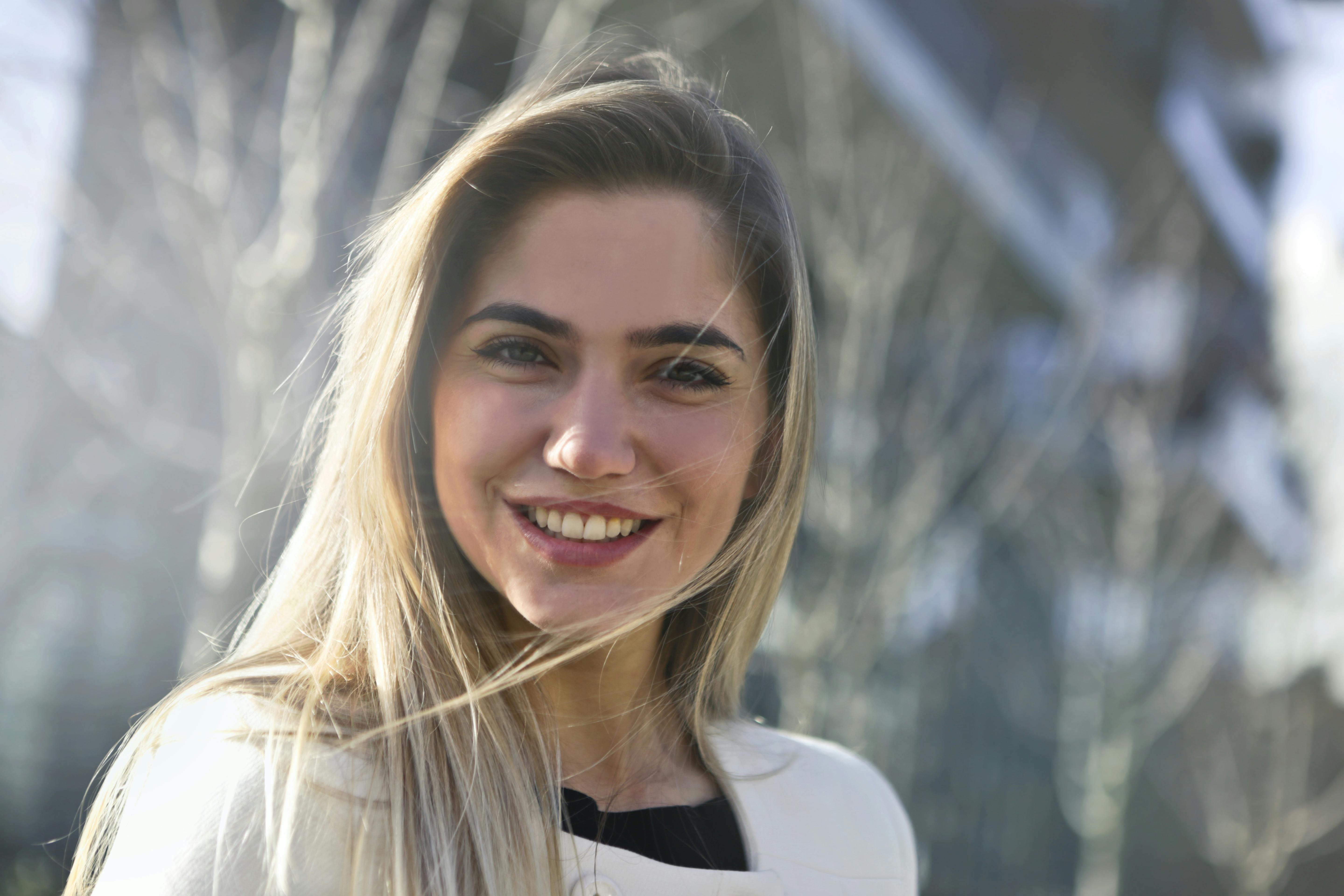 Smiling woman with long hair in a bright outdoor setting, sunlight reflecting off a modern building behind her.