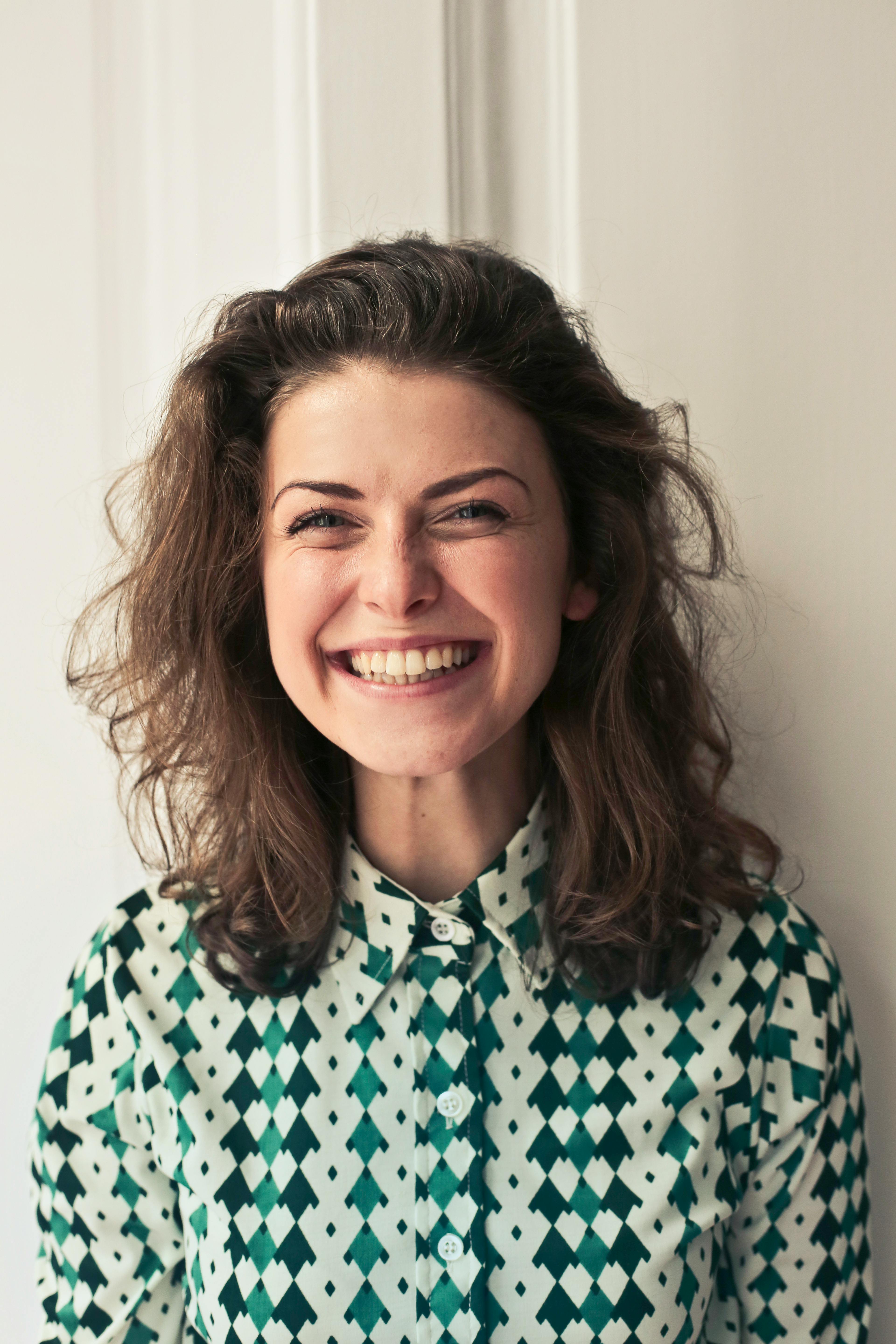 Smiling woman with brown curly hair wearing a green and white patterned collared shirt. She is very happy
