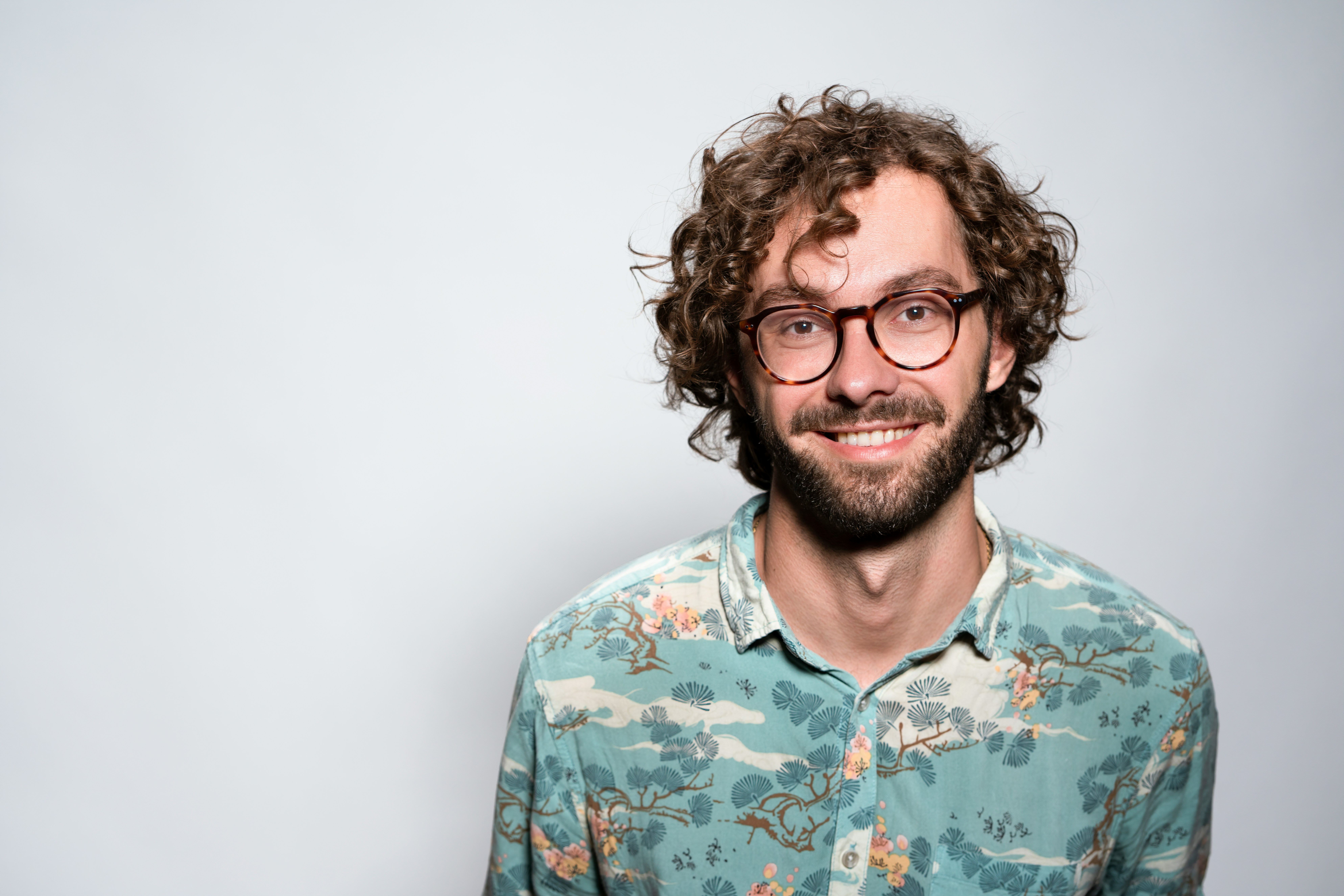 Happy man with curly hair, beard, and glasses smiles wearing a patterned shirt.